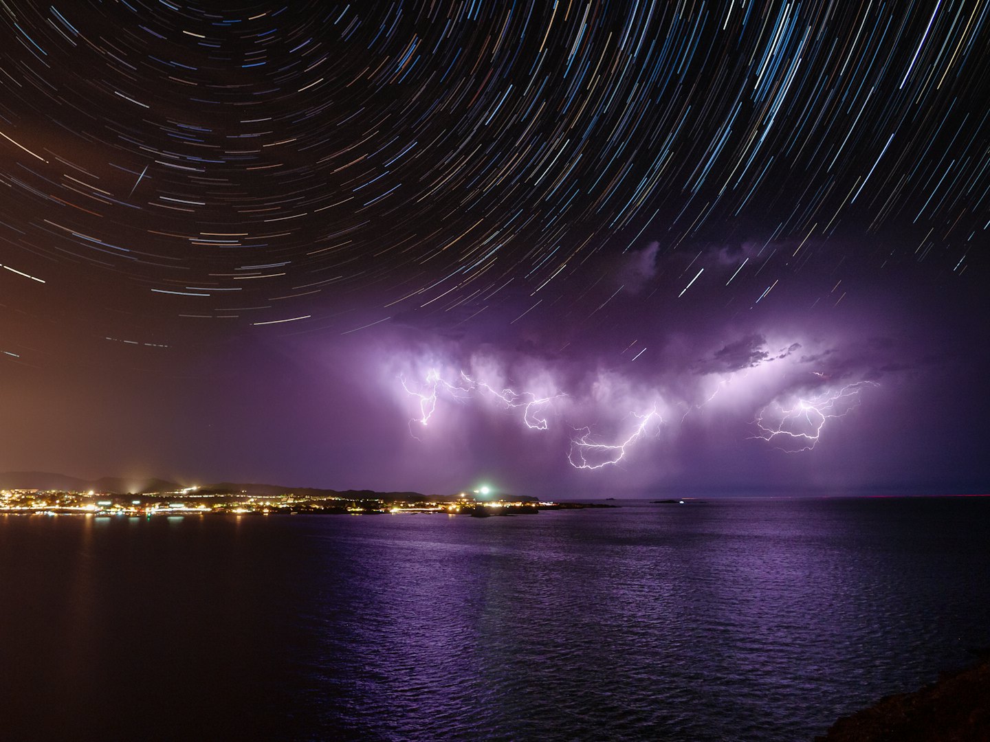 Star trails over a stormy sea with city lights.